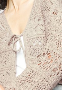Cárdigan de punto de encaje en beige claro con cierre de lazo, que presenta patrones intrincados y mangas abiertas, usado sobre una blusa blanca.