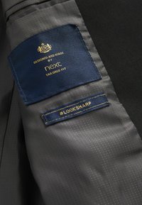 Etichetta in tessuto blu navy con scritte dorate che recitano "Progettato e realizzato da next tailored fit" accanto a un'etichetta grigia scura con la dicitura "#LOOKSHARP."