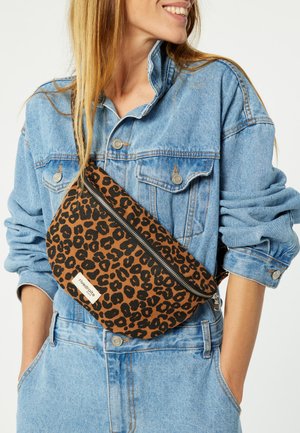 Braun gefleckte Umhängetasche aus Stoff mit Reißverschluss und einem Etikett. Die Tasche wird über einer Jeansjacke getragen.