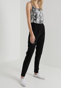 Svarta skräddarsydda byxor som matchas med en ärmlös topp i svart och vitt ormtryck. Modellen bär vita sneakers, vilket ger en avslappnad stil.