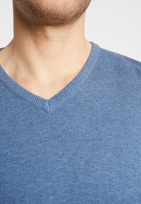 Niebieski sweter z dekoltem w serek, wykonany z dzianiny. Posiada prążkowany dekolt i gładką teksturę. Zbliżenie ukazujące szczegóły szycia.