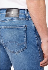Vue arrière d'une personne portant un jean en denim bleu avec une poche arrière et une étiquette de marque en cuir noir « Mavi » sur la ceinture.