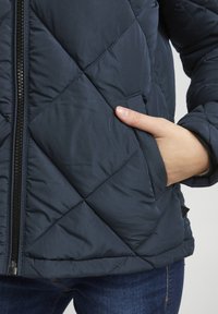 Marineblau gesteppte Jacke mit Reißverschluss, seitlichem Taschen, eleganter Textur und einem Saum, der auf Hüfthöhe sitzt.