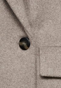 Tissu de manteau en mélange de laine gris avec une surface douce et texturée. Présente un bouton noir et un détail de poche passepoilée sur le côté avant.