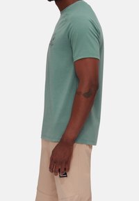 Camiseta de manga corta color verde menta, con cuello redondo, de tela suave, con estampado gráfico negro en el pecho, combinada con pantalones beige claros.