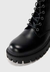 Bota de cuero negro con punta redonda, superficie texturizada, ojales metálicos y cordones negros, cuenta con una suela gruesa con estrías para mayor agarre.