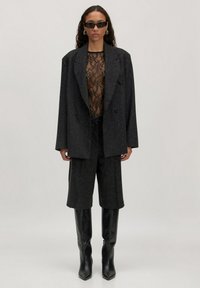 Vrouw in een oversized zwarte blazer, doorschijnende kanttop, zwarte shorts, kniehoog zwarte laarzen en donkere zonnebril die tegen een effen achtergrond staat.