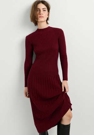 Frau trägt ein langärmliges, bordeauxrotes, geripptes Strickkleid, steht und hebt leicht den Rocksaum, schwarze kniehohe Stiefel sind sichtbar.