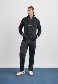 Juodas sportinis džemperis ir kelnės, pagamintos iš glotnaus audinio, su užtrauktuku, ant marškinėlių po juo yra žalias logotipas, o ant kojų baltos sportbačiai.