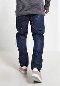 Jeans de mezclilla azul oscuro con un corte relajado, que cuentan con doble bolsillo trasero, costura amarilla y textura desgastada. Combinados con zapatillas beige.