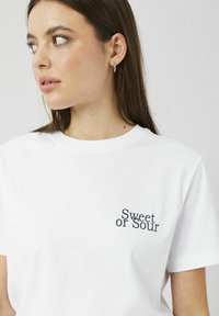 T-shirt blanc en coton avec manches courtes et col rond. Impression sur le côté gauche de la poitrine indiquant « Sweet or Sour » en police noire sans empattement.