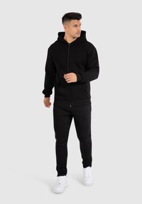 Zwarte zip-up hoodie met capuchon, gecombineerd met zwarte joggingsbroek. Inclusief witte sportschoenen. Zachte textuur, casual ontwerp.