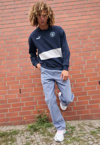Hanorac bleumarin cu dungi orizontale albe, logo Puma pe piept; asortat cu pantaloni de culoare albastră deschisă și adidași albi cu accente roz.