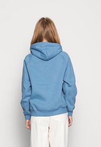 Persona con capelli lunghi che indossa un felpa blu e pantaloni bianchi, in piedi di fronte a uno sfondo bianco semplice con le mani ai lati.