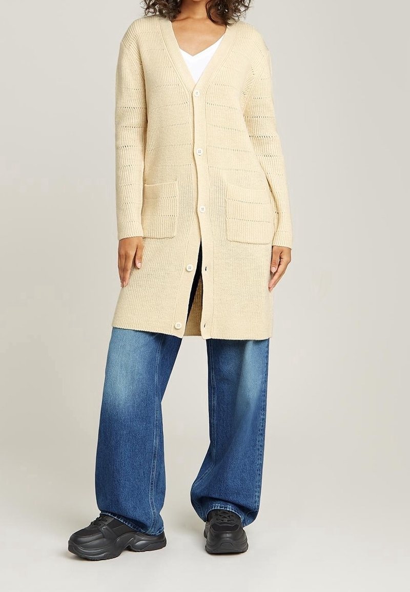 Personne portant un long cardigan tricoté crème avec des boutons, un haut blanc, un jean large bleu et des baskets noires, debout devant un fond gris.
