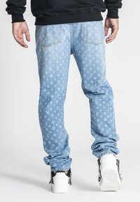 Jeans de denim azul claro con un patrón de logo en toda la prenda, forma ajustada y puños elásticos, combinados con zapatillas blancas y un suéter negro.