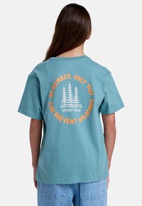 Camiseta de manga corta en color teal con un gráfico circular de árboles y texto en naranja y blanco en la parte posterior. Material de algodón; corte relajado.