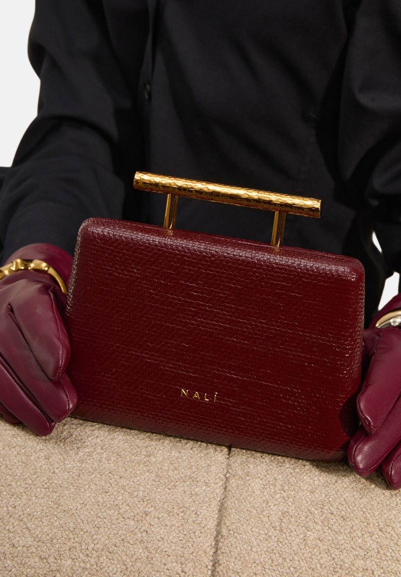 Borsa in pelle bordeaux con finitura testurizzata, dotata di manico in oro testurizzato e logo impresso, posizionata su una superficie beige.
