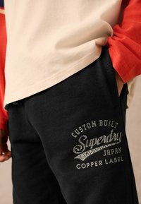 Schwarze Jogginghosen mit einem strukturierten Logo auf der Vorderseite, das "Superdry", "Custom Built Japan" und "Copper Label" zeigt. Sichtbar weicher Stoff.
