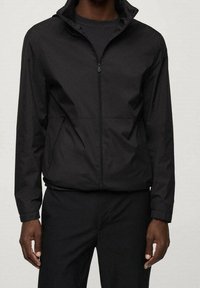 Veste légère noire avec un col haut, une fermeture éclair intégrale à l'avant et des poches latérales. Texture lisse et design minimaliste.