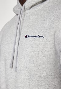 Jasnoszara bluza z kapturem wykonana z teksturowanej tkaniny, z przednią kieszenią typu kangur, kapturem ze sznurkiem oraz haftowanym logo Champion w granatowym i czerwonym kolorze.
