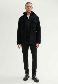 Granatowa kurtka z dużymi kieszeniami, nałożona na czarną pikowaną kamizelkę. Czarnie dopasowane spodnie i czarne botki za kostkę dopełniają całość.