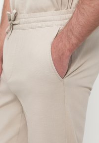 Μπεζ υφασμάτινο παντελόνι με ελαστική μέση, πλαϊνές τσέπες και απαλό ύφασμα. Διαθέτει ορατό κορδόνι για ρύθμιση.