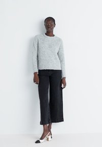Un pull en tricot gris duveteux associé à un jean large noir et des chaussures à talons blancs avec des accents noirs sur les orteils, le tout sur un fond neutre.