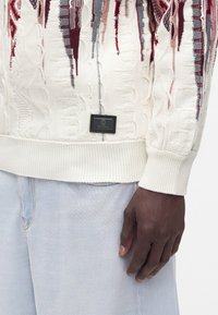 Gros plan d’une personne portant un pull texturé blanc avec des motifs rouges et gris et un jean en denim bleu clair.