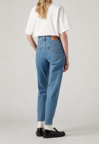 Femme debout, portant un jean bleu taille haute, un t-shirt blanc, des chaussettes blanches et des mocassins noirs, tournée de dos sur un fond uni.