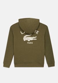 Φούτερ με κουκούλα σε ελιά πράσινο χρώμα, κατασκευασμένο από μαλακό ύφασμα, με μεγάλο λευκό γραφικό κροκόδειλου και το κείμενο "LACOSTE PARIS" στην πλάτη. Ρυθμιζόμενη κουκούλα.