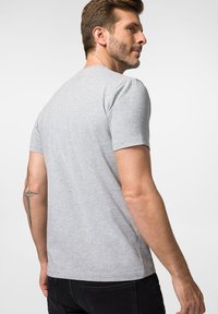 Szara koszulka z krótkim rękawem, uszyta z miękkiego materiału, z klasycznym okrągłym dekoltem i luźnym krojem, widoczna z tyłu z tatuażem na ramieniu.