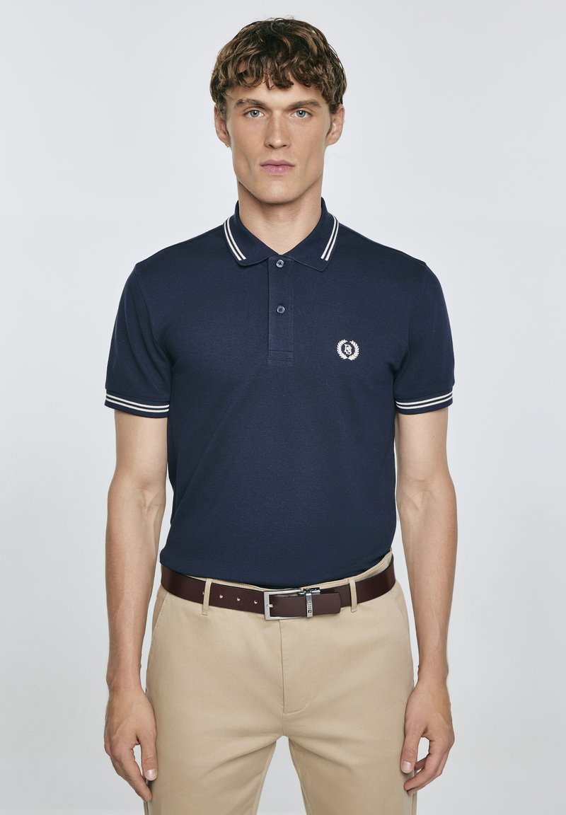 Tamsiai mėlyna polo maikė su dviem baltomis juostelėmis ant apykaklės ir rankovių, su nedideliu emblema kairėje krūtinės pusėje. Dėvima su smėlio spalvos kelnėmis ir rudomis diržais.