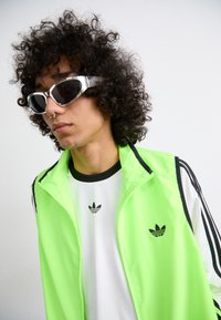 Neon zöld, cipzáros dzseki fekete részletekkel, fehér póló felett, amelyen egy kicsi fekete logó van. Négyzet alakú napszemüveg sötét lencsékkel.