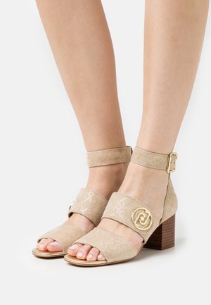 Sandalias de tacón de ante beige con correa en el tobillo, diseño de punta abierta, patrón en relieve y detalle metálico dorado con un logo circular.