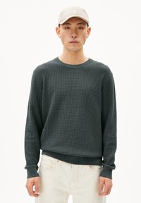 Pull en maille tissée vert foncé avec des manches longues et un col rond, associé à un jean clair. La mannequin porte une casquette beige clair.