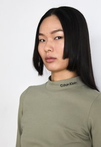 Olive green mock neck top made of smooth fabric, featuring a black 'Calvin Klein' logo at the collar, with a fitted design.