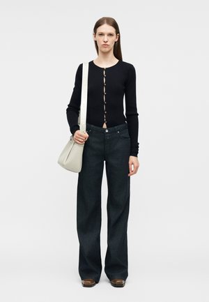 Femme portant un haut noir à manches longues boutonné, un jean large foncé, des chaussures marron, et portant un sac à bandoulière gris clair avec une sangle blanche.