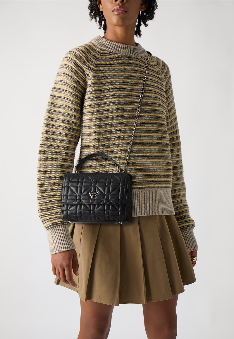 Schwarze gesteppte Handtasche mit einem oberen Griff und einer Kettenriemen, versehen mit einem dreieckigen Logo. Kombiniert mit einem gestreiften Pullover und einem plissierten Rock.