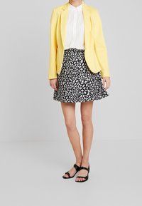 Blazer jaune porté sur une chemise blanche, assorti d'une jupe noire avec un motif floral blanc. Des sandales noires à fines brides complètent la tenue.