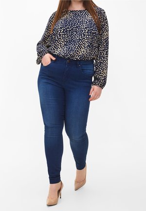 Femme portant un jean skinny bleu foncé, une chemise bleu marine avec des taches claires, des talons hauts beige, debout avec une main dans la poche.