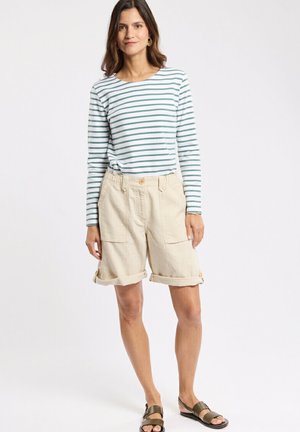Femme portant un t-shirt à manches longues rayé vert et blanc, un short beige retroussé avec des poches, et des sandales vert foncé, debout sur un fond blanc.