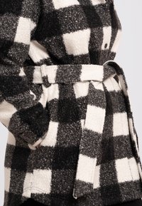 Černobílá kostkovaná fleecová bunda s texturovaným povrchem, páskem v pase a bočními kapsami, má velké knoflíky na přední straně.