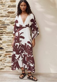 Maxikleid in Weiß mit kastanienbraunen Blattmustern, V-Ausschnitt und ausgestellten Ärmeln. Das Kleid hat mehrlagige Rüschen am Saum und wird mit schwarzen Sandalen mit Riemchen getragen.