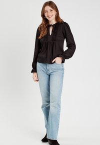 Blouse noire à manches longues avec un col en trou de serrure, détails en dentelle et poignets froncés, associée à un jeans droit bleu clair et des bottines noires.