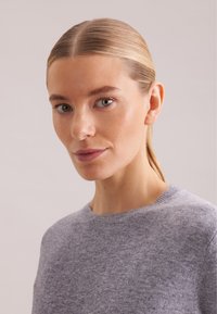 Grauer Strickpullover mit rundem Ausschnitt, aus weichem Material, mit einem strukturierten Muster. Modell mit glattem Haar, neutraler Hintergrund.