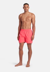 Giovane uomo che sta in piedi a piedi nudi, indossando pantaloni da bagno corallo luminosi contro uno sfondo bianco semplice.