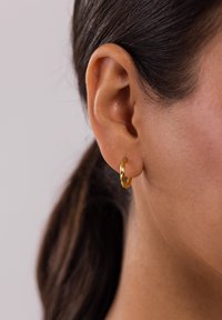 Goldene Creole, gewundenes Design, kreisförmige Form, reflektierende Textur, am Ohr präsentiert vor einem neutralen Hintergrund.