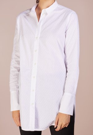 Kobieta ubrana w długą białą koszulę zapinaną na guziki o subtelnej kropkowanej fakturze i kontrastujących gładkich białych mankietach, w zestawieniu z czarnymi spodniami.