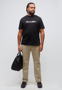 Homme aux cheveux tressés portant un t-shirt graphique noir, un pantalon beige, des chaussures noires, tenant un sac de voyage en cuir noir, debout devant un fond blanc.